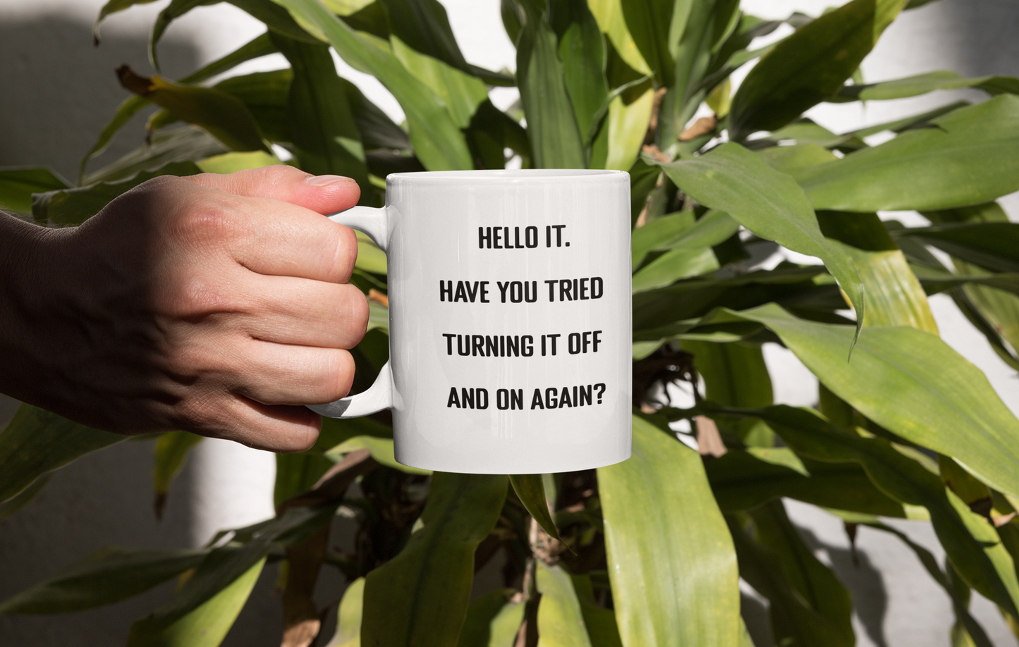 A glossy white 11oz mug with the words 'Hello IT. Have you tried turning it off and on again?' printed in the centre of the mug in simple black block font. A hand is holding the mug up in front of a large house plant with lots of green leaves.