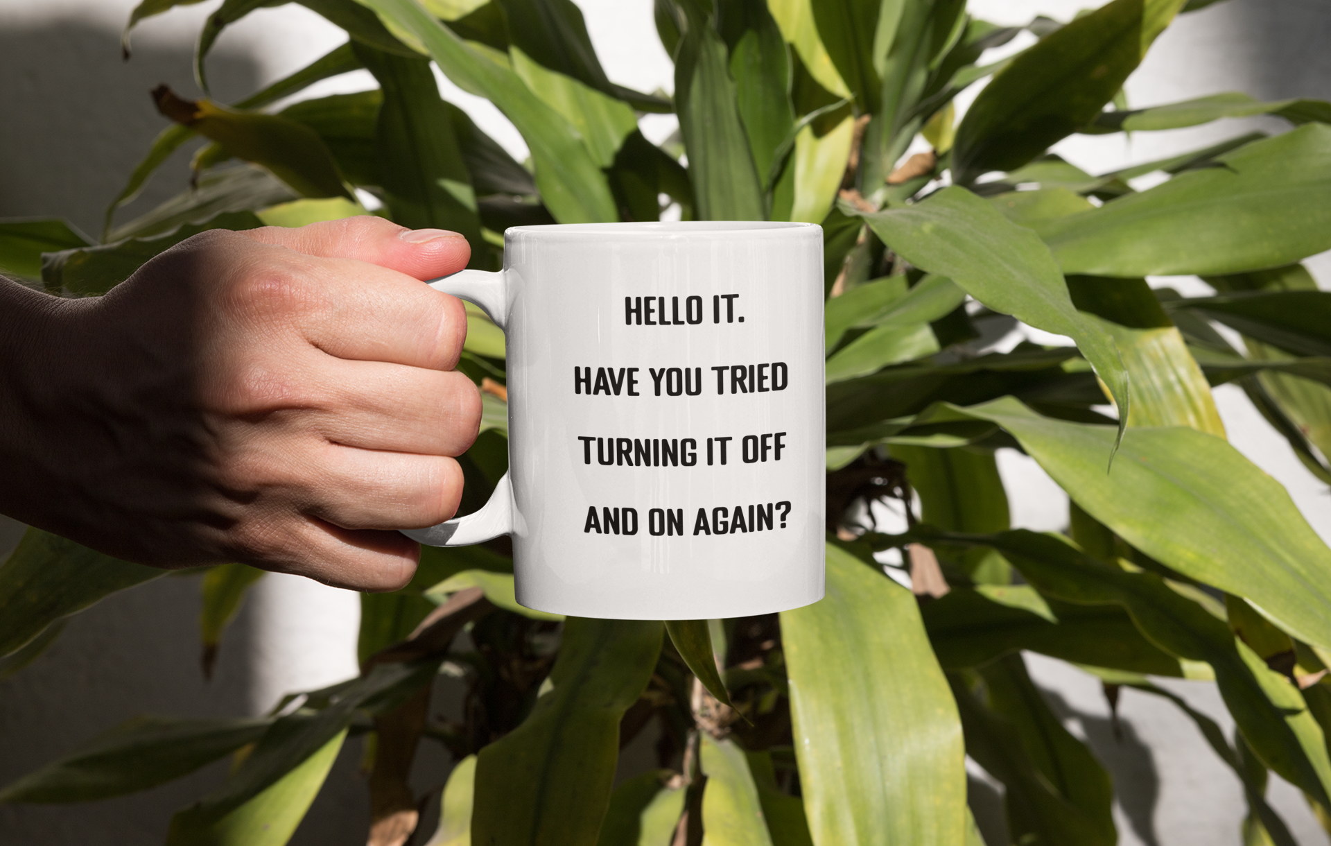 A glossy white 11oz mug with the words 'Hello IT. Have you tried turning it off and on again?' printed in the centre of the mug in simple black block font. A hand is holding the mug up in front of a large house plant with lots of green leaves. 