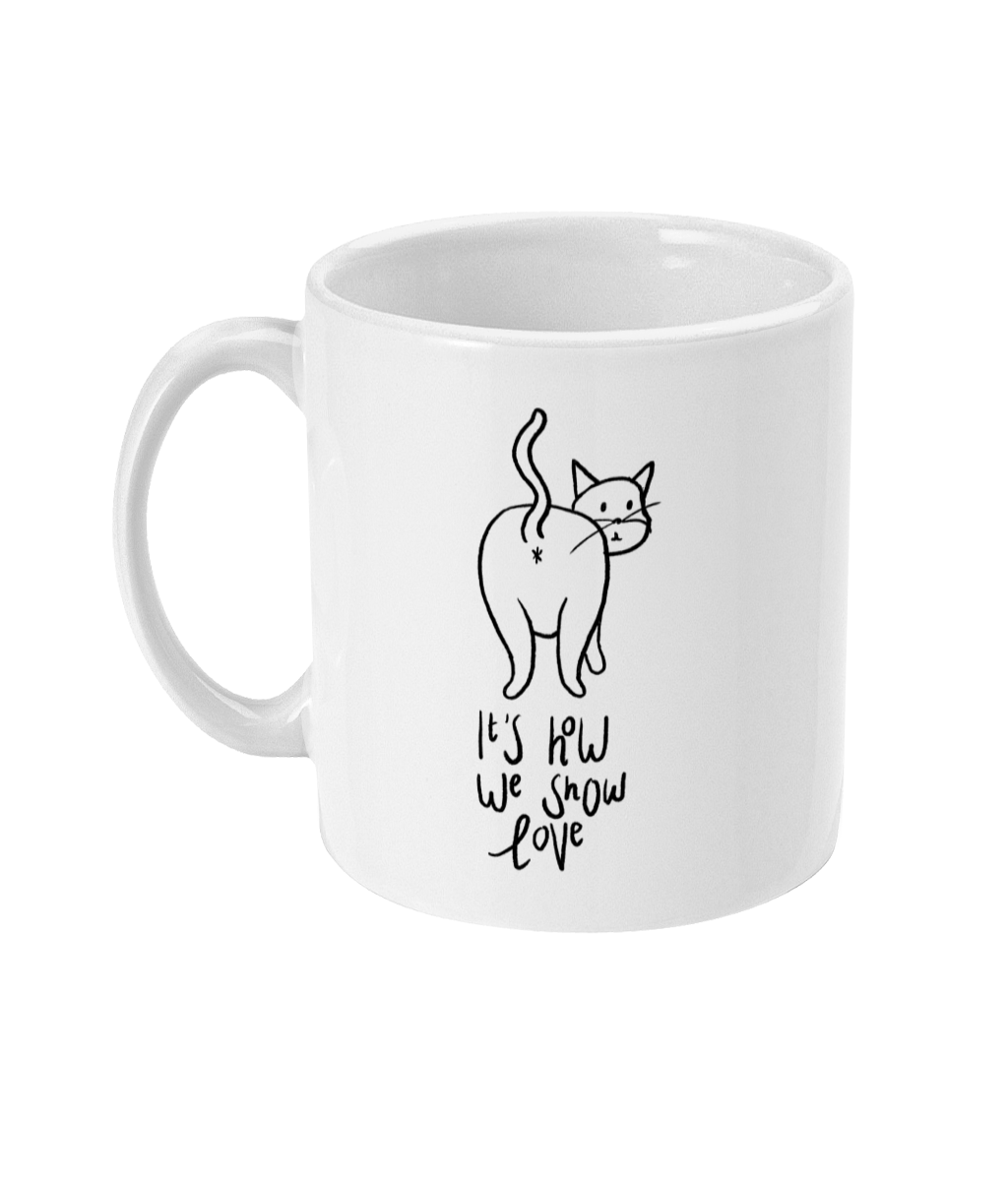 A 11oz glossy white mug with a cartoon drawn cat showing it's butthole for everyone to see. Underneath the cartoon is the saying 'It's how we show love' in handwritten cursive black font.  The mug is placed against a white background.