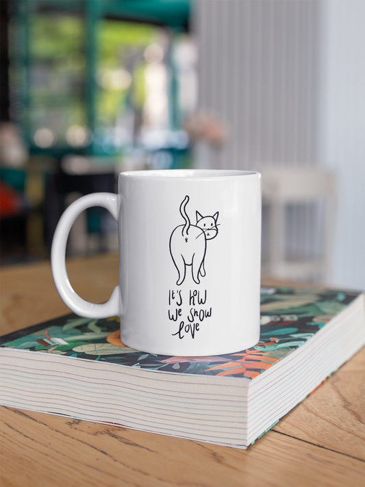 A 11oz glossy white mug with a cartoon drawn cat showing it's butthole for everyone to see. Underneath the cartoon is the saying 'It's how we show love' in handwritten cursive black font. The mug is placed on a large colourful book. 