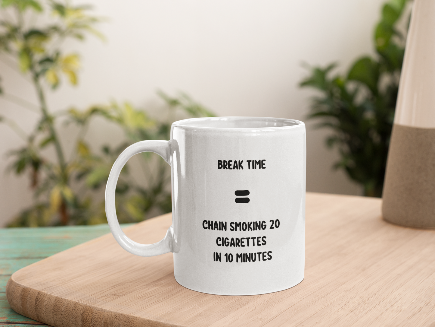 A 11oz glossy white mug with the quote 'Break time = Chain smoking 20 cigarettes in 10 minutes' in black bold font placed in its centre. The mug is placed on a chopping board with two large plants blurred in the background. 