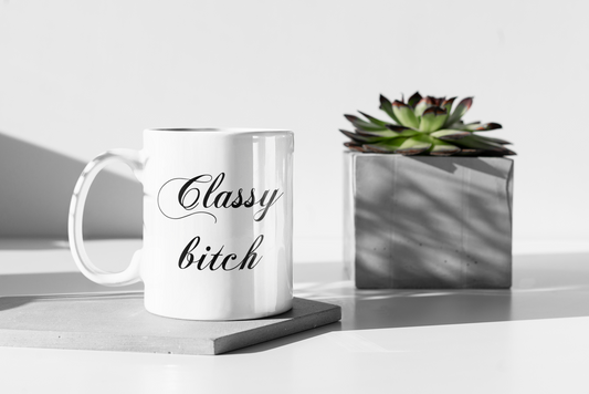 A 11oz glossy white mug with the words 'Classy bitch' in large cursive black font placed in its centre. The mug is place on a grey hexagon place mat and is next to a succulent plant in a grey square pot.   
