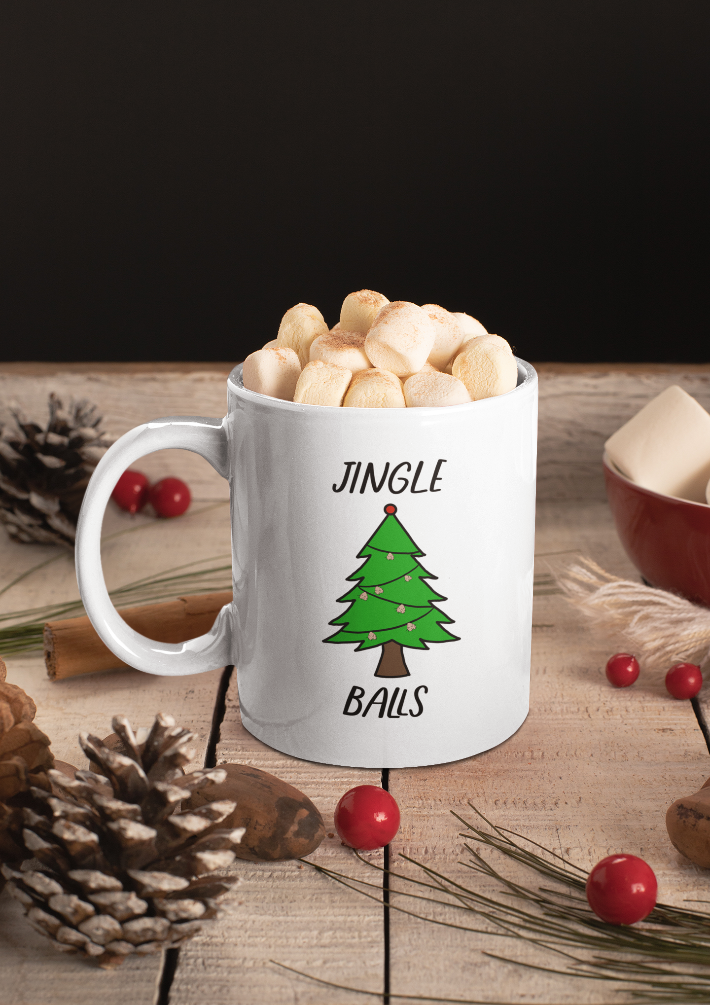 A glossy white 11oz mug with a cartoon drawn green Christmas tree with string ball sack lights circling the tree. The word Jingle is printed above the tree in black font and the word Balls is printed below the tree in black font. There are marsh mellows in the mug and the mug is surrounded by festive pinecones and berries. 