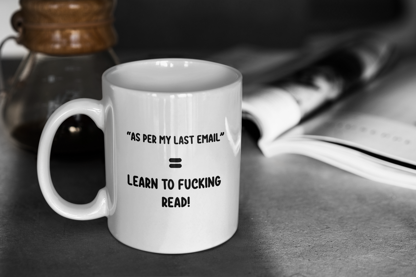 A glossy white 11oz mug with the statement 'As per my last email =  Learn to fucking read!' Placed upon it's centre. Behind the mug can be seen a blurred image of a coffee pot and magazine. 