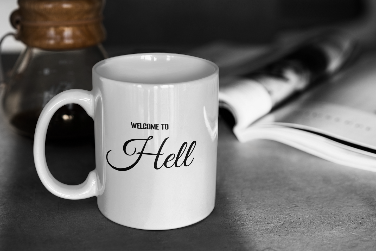 A glossy white 11oz mug with the words 'Welcome to Hell' Printed in the centre in block and cursive font. The mug is placed on a grey table top and a glass coffee pot and grey magazine can be seen in the background slightly blurred.